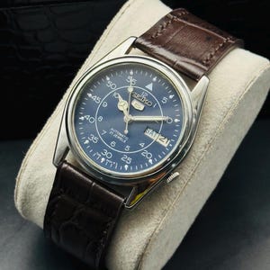 May include: A stainless steel Seiko automatic watch with a blue dial and a brown leather strap. The watch face displays the day and date, with white numerals and hands. The watch is displayed on a cream-colored cushion.