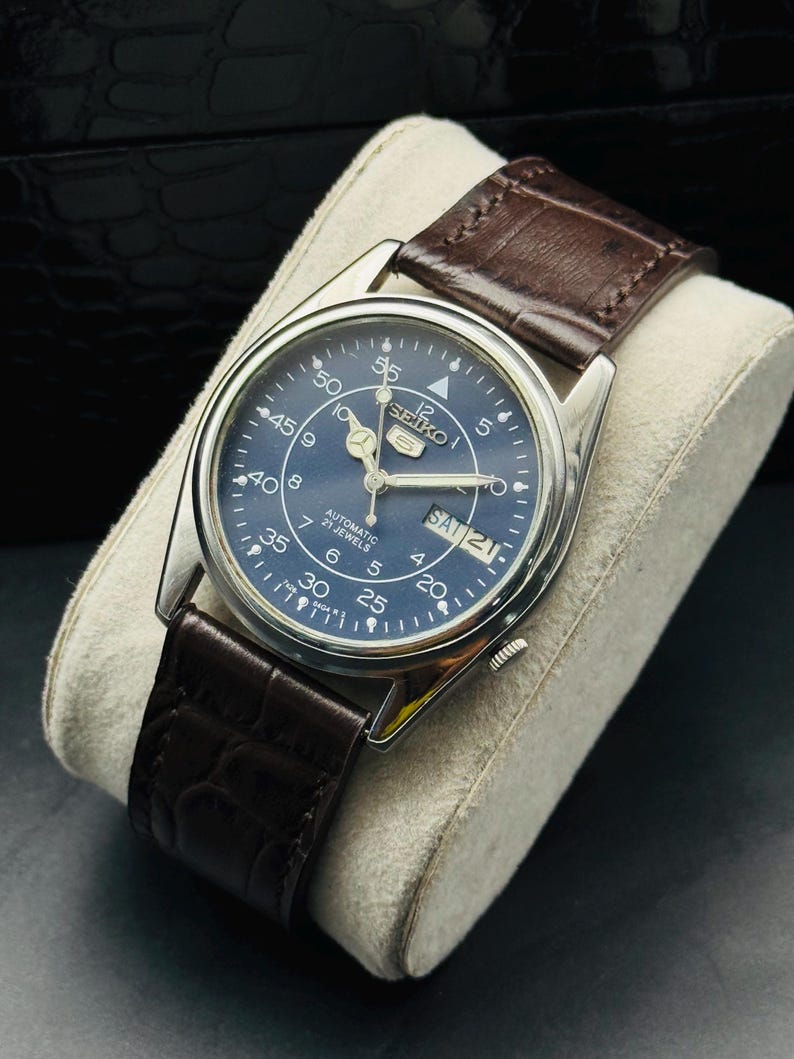May include: A stainless steel Seiko automatic watch with a blue dial and a brown leather strap. The watch face displays the day and date, with white numerals and hands. The watch is displayed on a cream-colored cushion.