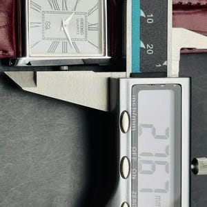 May include: A rectangular Seiko Quartz watch with a silver face, Roman numerals, and a burgundy leather band. The watch is being measured with a digital caliper, showing a measurement of 27.67 mm.