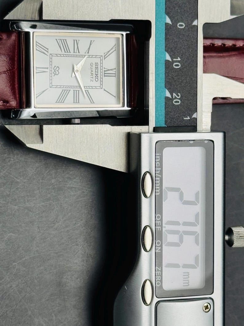 May include: A rectangular Seiko Quartz watch with a silver face, Roman numerals, and a burgundy leather band. The watch is being measured with a digital caliper, showing a measurement of 27.67 mm.
