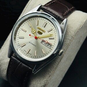 May include: Silver-toned Seiko automatic watch with a light silver dial, gold hands, and a red second hand. The watch features a brown leather strap and a date display. The watch is marked "Japan Made".