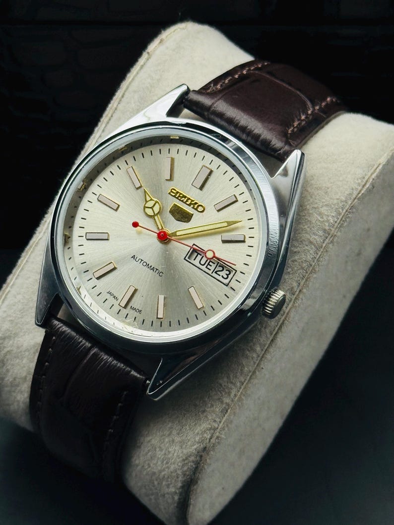 May include: Silver-toned Seiko automatic watch with a light silver dial, gold hands, and a red second hand. The watch features a brown leather strap and a date display. The watch is marked "Japan Made".