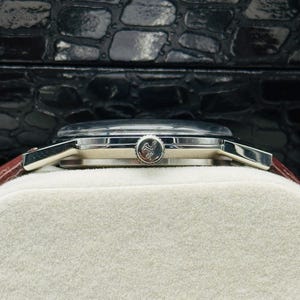 May include: A silver wristwatch with a brown leather band. The watch face is silver with a small emblem. The watch is resting on a white surface, with a black textured background.