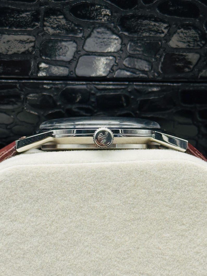 May include: A silver wristwatch with a brown leather band. The watch face is silver with a small emblem. The watch is resting on a white surface, with a black textured background.