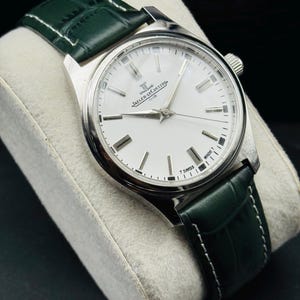 May include: A silver-toned Jaeger-LeCoultre Club watch with a white face and green leather band. The watch has a round face with silver hour markers and hands. The green band is stitched with white thread.
