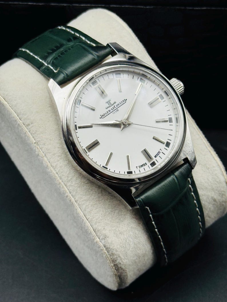 May include: A silver-toned Jaeger-LeCoultre Club watch with a white face and green leather band. The watch has a round face with silver hour markers and hands. The green band is stitched with white thread.