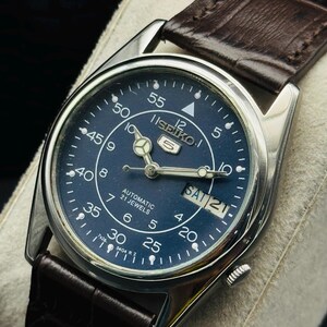 May include: A silver-toned Seiko 5 automatic watch with a dark blue face and brown leather strap. The watch face displays the day and date, with white numerals and hands. The strap is a dark brown, textured leather.