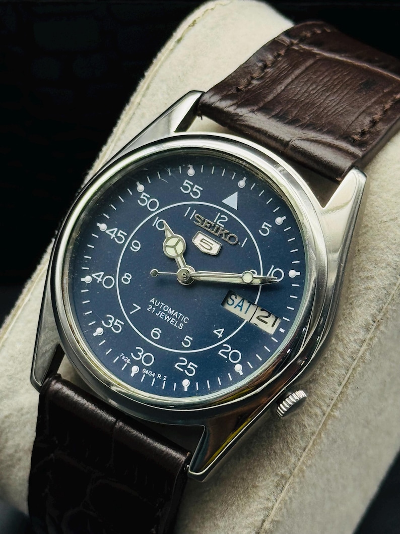 May include: A silver-toned Seiko 5 automatic watch with a dark blue face and brown leather strap. The watch face displays the day and date, with white numerals and hands. The strap is a dark brown, textured leather.