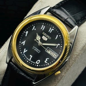Op de afbeelding: Een Seiko 5 automatisch polshorloge met een zwarte wijzerplaat, gouden bezel en Arabische cijfers. Het horloge heeft een dag-datumweergave en een zwarte leren band. De wijzerplaat heeft ook de tekst "AUTOMATIC 21 JEWELS" en "MADE IN JAPAN".