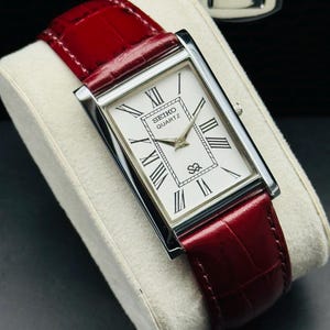 May include: A rectangular Seiko Quartz watch with a white face and black Roman numerals. The watch has a silver case and a red leather band. The watch is displayed on a white cushion.