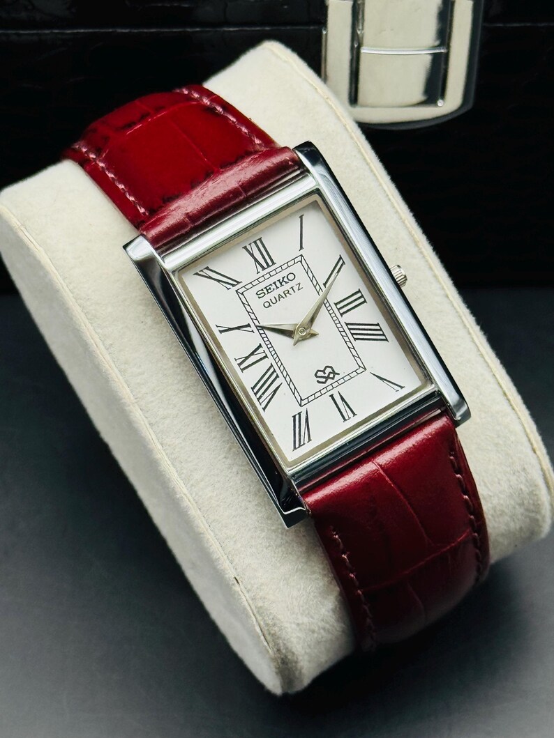 May include: A rectangular Seiko Quartz watch with a white face and black Roman numerals. The watch has a silver case and a red leather band. The watch is displayed on a white cushion.
