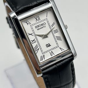 Puede incluir: Reloj Seiko Quartz rectangular con esfera blanca, n&uacute;meros romanos negros y correa de cuero negra. El reloj tiene una caja plateada y una correa de cuero negro texturizada. La esfera muestra el logotipo de Seiko Quartz.