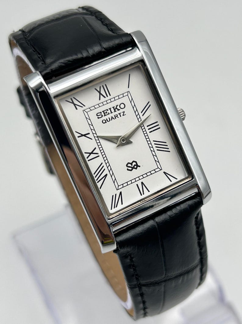 Puede incluir: Reloj Seiko Quartz rectangular con esfera blanca, n&uacute;meros romanos negros y correa de cuero negra. El reloj tiene una caja plateada y una correa de cuero negro texturizada. La esfera muestra el logotipo de Seiko Quartz.