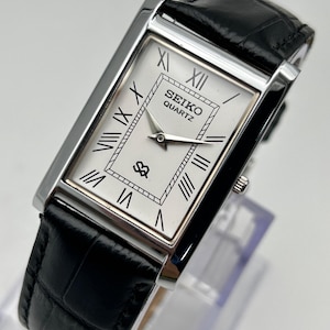 Könnte beinhalten: Eine rechteckige Seiko Quartz-Uhr mit silberfarbenem Gehäuse und weißem Zifferblatt mit schwarzen römischen Ziffern. Die Uhr hat ein schwarzes Lederarmband mit Krokodilmuster. Das Zifferblatt zeigt das Seiko Quartz-Logo.