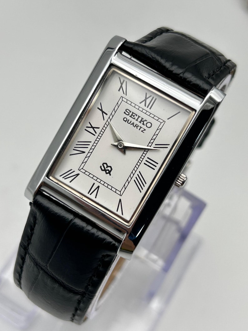 Puede incluir: Un reloj Seiko Quartz rectangular con caja plateada y esfera blanca con n&uacute;meros romanos negros. El reloj tiene una correa de cuero negro con estampado de cocodrilo. La esfera muestra el logotipo de Seiko Quartz.