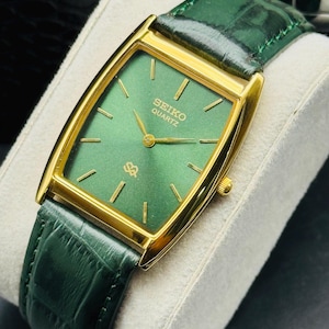 May include: A rectangular Seiko Quartz watch with a gold-tone case and a green dial. The watch features gold-tone hands and markers, and a green leather strap. The watch is displayed on a white cushion.