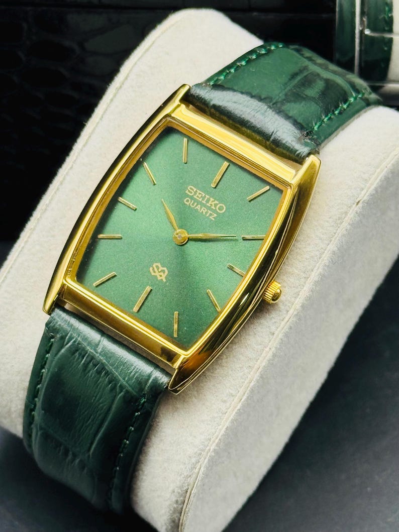 May include: A rectangular Seiko Quartz watch with a gold-tone case and a green dial. The watch features gold-tone hands and markers, and a green leather strap. The watch is displayed on a white cushion.