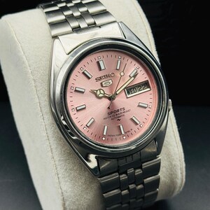 May include: A stainless steel Seiko 5 Sports automatic watch with a pink dial. The watch features a day and date display and a silver-tone bracelet. The watch is displayed on a white cushion.