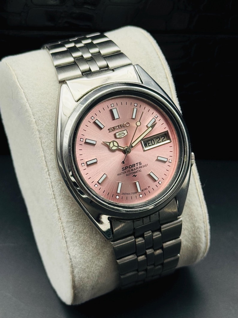 May include: A stainless steel Seiko 5 Sports automatic watch with a pink dial. The watch features a day and date display and a silver-tone bracelet. The watch is displayed on a white cushion.