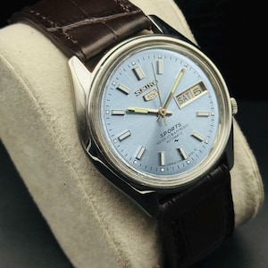 May include: A silver-toned Seiko 5 Sports automatic watch with a light blue face and a brown leather strap. The watch face displays the day and date, with the day reading "SAT 5". The watch has a classic design.