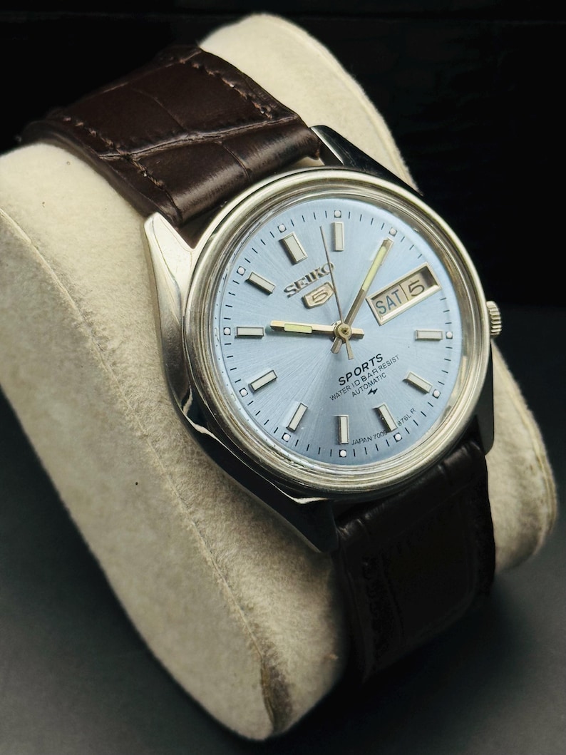 May include: A silver-toned Seiko 5 Sports automatic watch with a light blue face and a brown leather strap. The watch face displays the day and date, with the day reading "SAT 5". The watch has a classic design.
