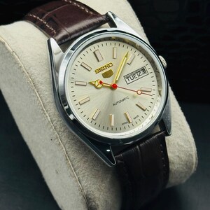 May include: A silver-toned Seiko automatic wristwatch with a light-colored face and a brown leather band. The watch face displays the day and date, with gold-colored hands and markers. The band is a dark brown with a textured pattern.