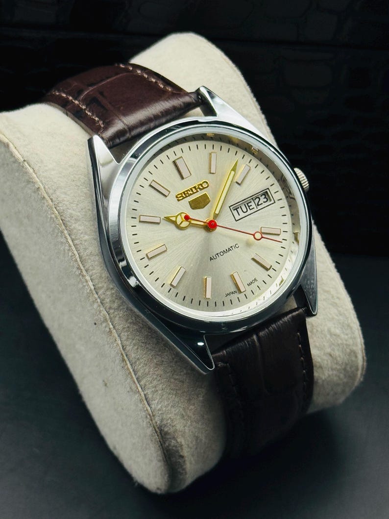 May include: A silver-toned Seiko automatic wristwatch with a light-colored face and a brown leather band. The watch face displays the day and date, with gold-colored hands and markers. The band is a dark brown with a textured pattern.
