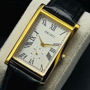 May include: A rectangular gold-toned Seiko watch with a white face, black Roman numerals, and a small seconds dial. The watch has gold hands and a black leather band. The watch is displayed on a white cushion.