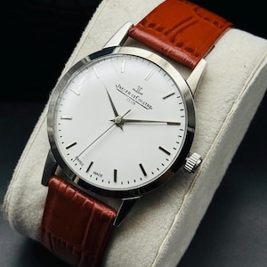 Puede incluir: Un reloj Jaeger-LeCoultre Club plateado con esfera blanca, manecillas y marcadores negros. La correa es de cuero marr&oacute;n. El reloj se exhibe sobre un coj&iacute;n gris.