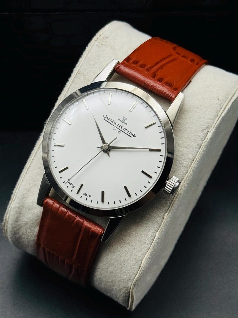 Puede incluir: Un reloj Jaeger-LeCoultre Club plateado con esfera blanca, manecillas y marcadores negros. La correa es de cuero marr&oacute;n. El reloj se exhibe sobre un coj&iacute;n gris.