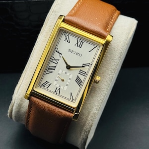 May include: A rectangular gold-toned Seiko watch with a light-coloured face and black Roman numerals. The watch has a brown leather strap. The watch is displayed on a light-coloured cushion.