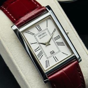 May include: A rectangular Seiko Quartz watch with a white face, black Roman numerals, and silver hands. The watch has a silver case and a burgundy leather band. The watch is displayed on a white surface.