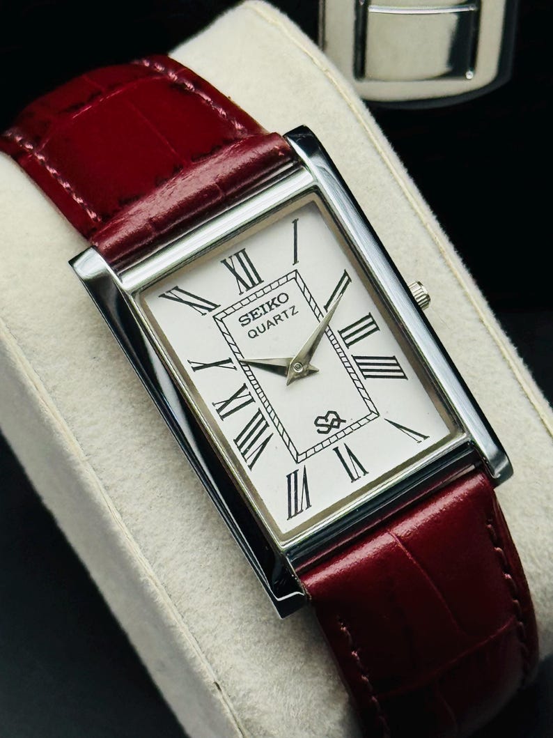 May include: A rectangular Seiko Quartz watch with a white face, black Roman numerals, and silver hands. The watch has a silver case and a burgundy leather band. The watch is displayed on a white surface.