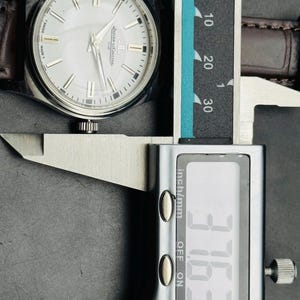 May include: A silver-toned wristwatch with a white face and brown leather band is measured with a digital caliper. The watch face has silver markers and the brand name "Jaeger-LeCoultre". The caliper reads 37.63 mm.