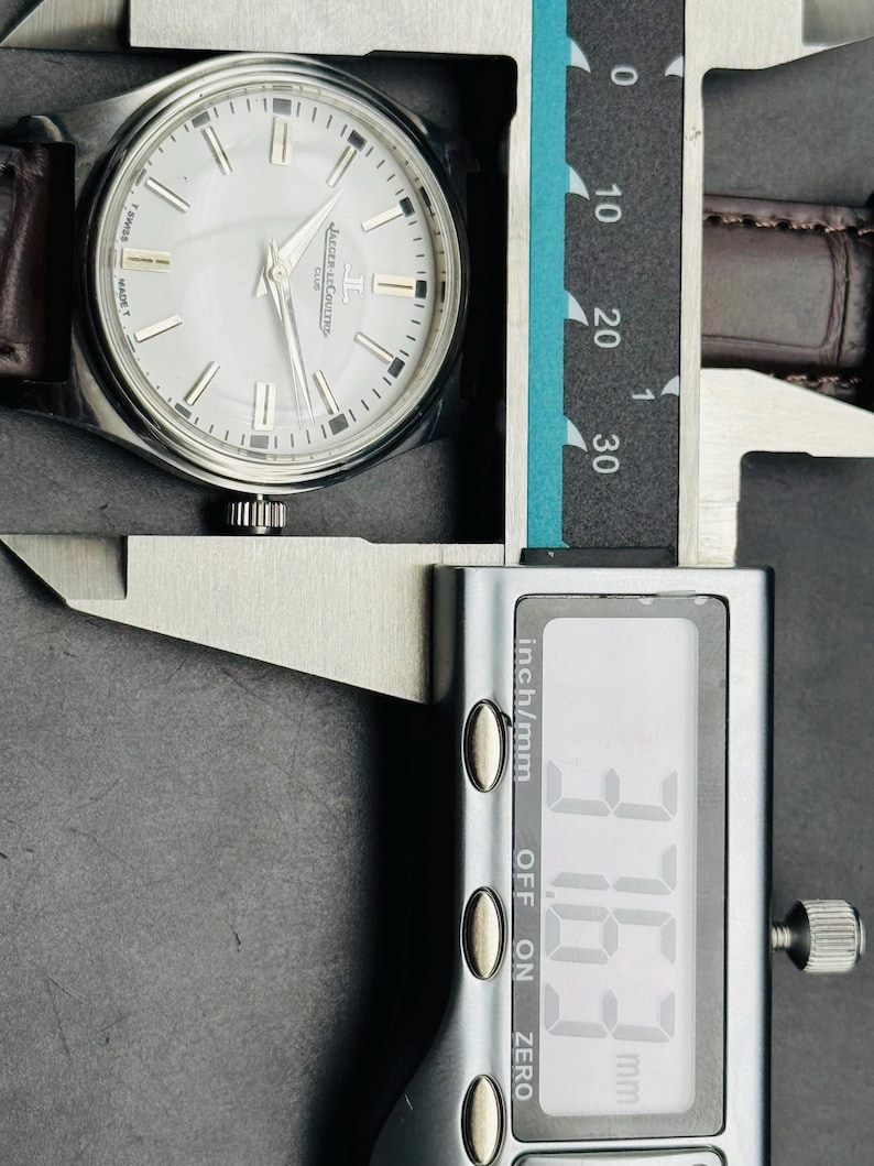 May include: A silver-toned wristwatch with a white face and brown leather band is measured with a digital caliper. The watch face has silver markers and the brand name "Jaeger-LeCoultre". The caliper reads 37.63 mm.