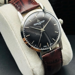 May include: A silver-toned Jaeger-LeCoultre Club watch with a dark face and brown leather band. The watch has a round face with silver hands and markers. The band is a rich brown color and appears to be made of leather. The watch is displayed on a white cushion.