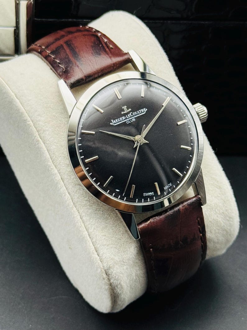 May include: A silver-toned Jaeger-LeCoultre Club watch with a dark face and brown leather band. The watch has a round face with silver hands and markers. The band is a rich brown color and appears to be made of leather. The watch is displayed on a white cushion.