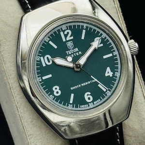 May include: A vintage Tudor watch with a dark green face, white hands, and a silver case. The watch has a black leather strap and the text "Tudor" and "Shock Resisting" are visible. The watch is displayed on a white surface.