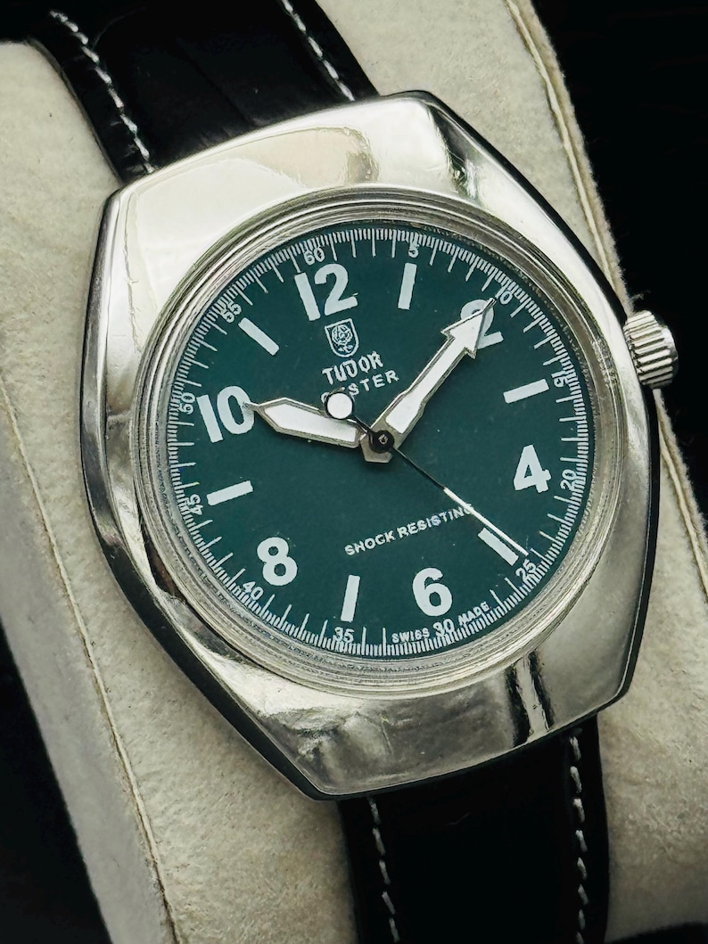 May include: A vintage Tudor watch with a dark green face, white hands, and a silver case. The watch has a black leather strap and the text "Tudor" and "Shock Resisting" are visible. The watch is displayed on a white surface.
