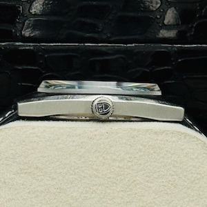 May include: A close-up of a silver wristwatch with a rectangular face and a black leather strap. The watch has a small, circular emblem on the face and is set against a cream-coloured background. The watch is in a black box.