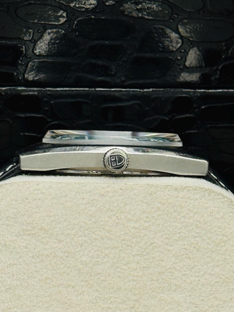 May include: A close-up of a silver wristwatch with a rectangular face and a black leather strap. The watch has a small, circular emblem on the face and is set against a cream-coloured background. The watch is in a black box.