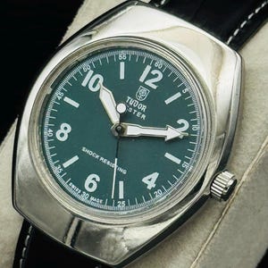 May include: A classic wristwatch featuring a green dial, silver casing, and a black leather strap. The watch face displays the brand name "TUDOR" and the text "SHOCK RESISTING". The watch has white hands and hour markers.