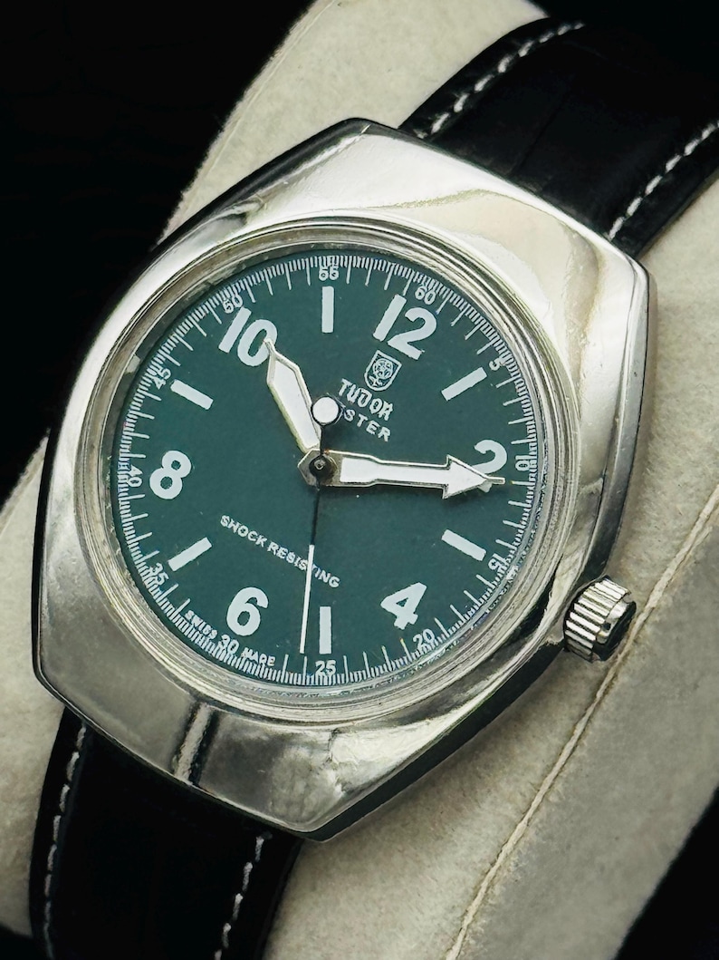 May include: A classic wristwatch featuring a green dial, silver casing, and a black leather strap. The watch face displays the brand name "TUDOR" and the text "SHOCK RESISTING". The watch has white hands and hour markers.