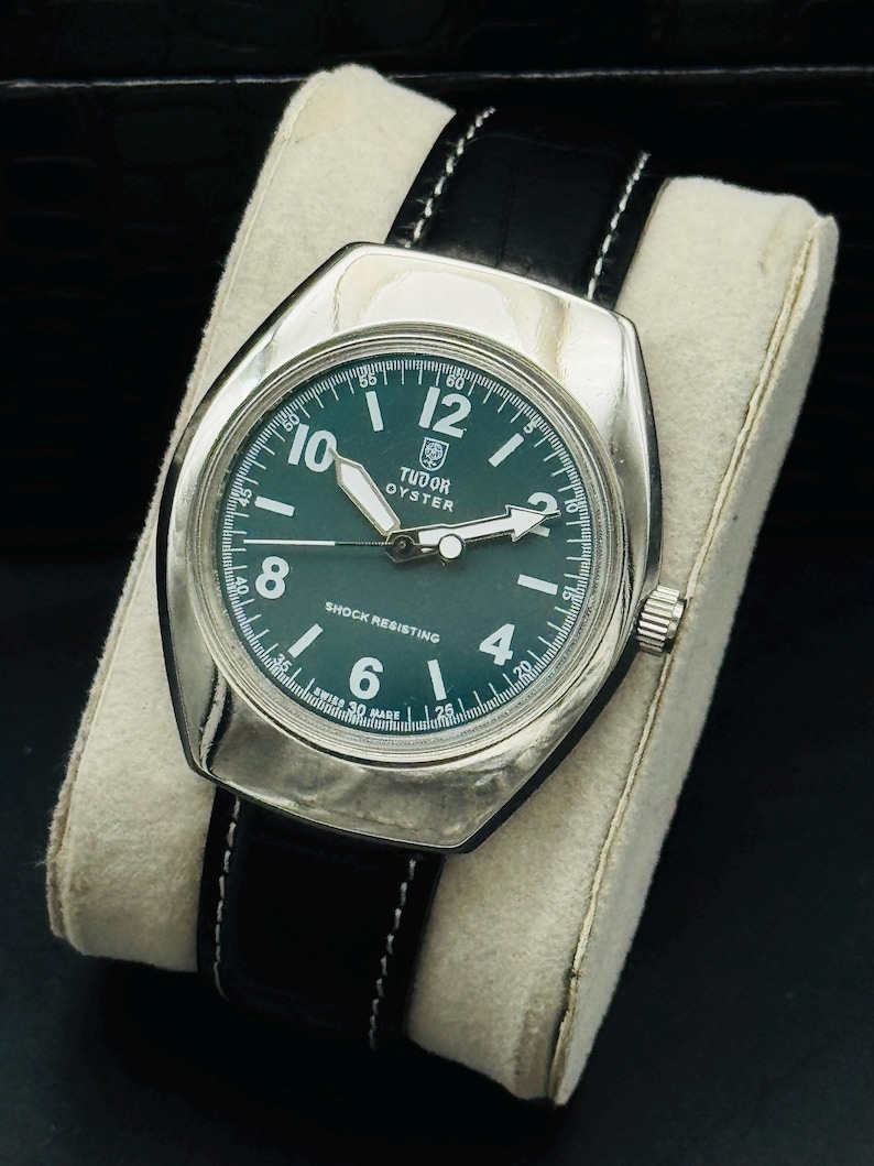 May include: A vintage Tudor Oyster watch with a silver case and a dark green dial. The watch features white hands and hour markers, a black leather strap with white stitching, and the text "SHOCK RESISTING".
