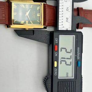 Puede incluir: Un reloj Seiko Quartz dorado con esfera verde y n&uacute;meros romanos. Tiene una correa de cuero marr&oacute;n y se mide con un calibrador digital. El calibrador muestra 27,2 mm. La esfera del reloj dice 'SEIKO Quartz'.