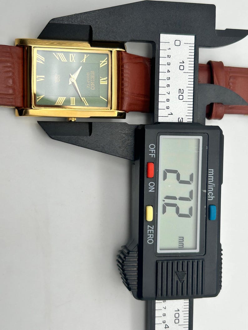 Puede incluir: Un reloj Seiko Quartz dorado con esfera verde y n&uacute;meros romanos. Tiene una correa de cuero marr&oacute;n y se mide con un calibrador digital. El calibrador muestra 27,2 mm. La esfera del reloj dice 'SEIKO Quartz'.