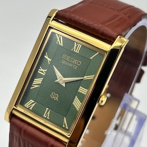 Puede incluir: Un reloj Seiko Quartz rectangular con caja dorada y esfera verde con n&uacute;meros romanos. El reloj tiene una correa de cuero marr&oacute;n. La esfera del reloj muestra el logotipo de Seiko Quartz.