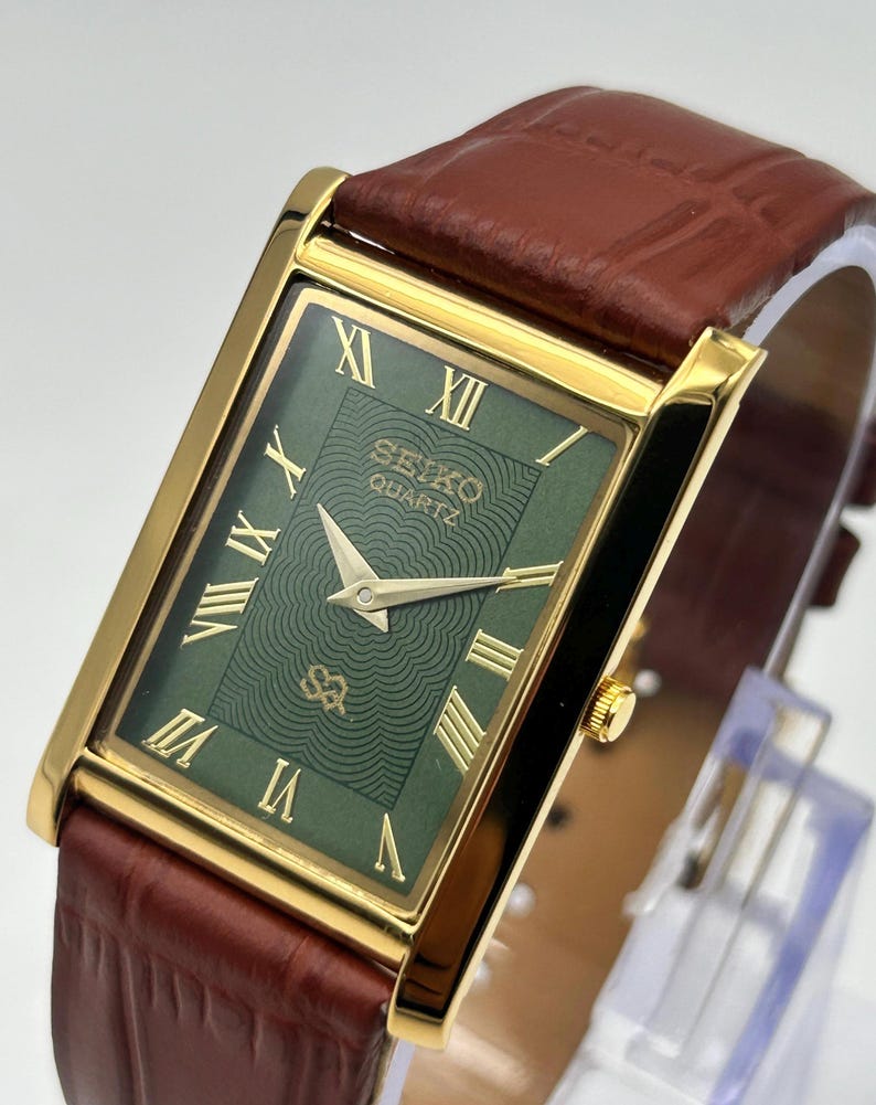 Puede incluir: Un reloj Seiko Quartz rectangular con caja dorada y esfera verde con n&uacute;meros romanos. El reloj tiene una correa de cuero marr&oacute;n. La esfera del reloj muestra el logotipo de Seiko Quartz.