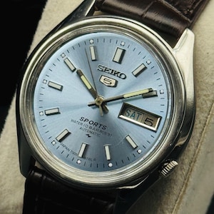 May include: A silver-toned Seiko 5 Sports watch with a light blue dial and a brown leather strap. The watch face displays the day and date, with the day reading "SAT 5". The watch has the text "SPORTS WATER 10 BAR RESIST AUTOMATIC" on the dial.