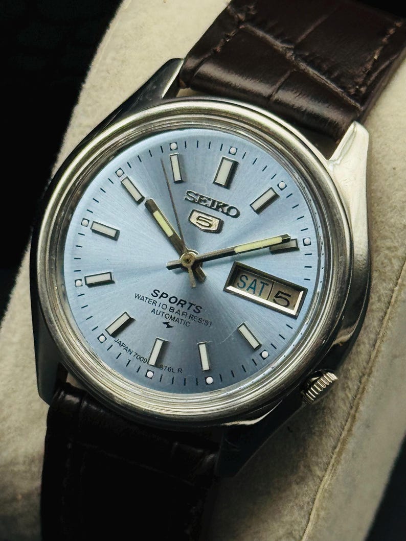May include: A silver-toned Seiko 5 Sports watch with a light blue dial and a brown leather strap. The watch face displays the day and date, with the day reading "SAT 5". The watch has the text "SPORTS WATER 10 BAR RESIST AUTOMATIC" on the dial.